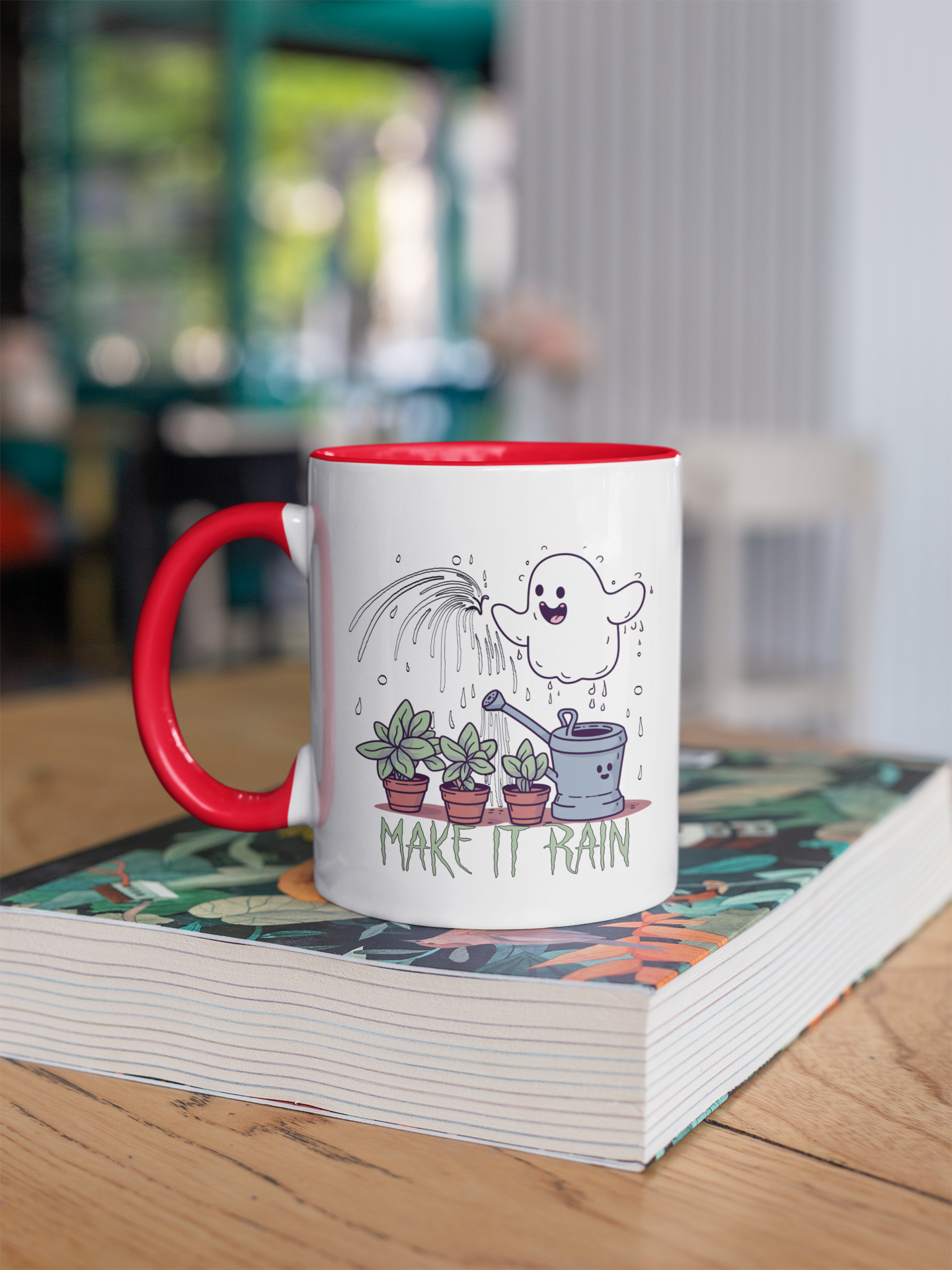 The photo displays a two-toned mug with a red handle and a white body, resting on a hardcover book on a wooden surface. The mug features a playful design with a cheerful ghost-like figure showering water over three potted plants, accompanied by a watering can and the phrase "MAKE IT RAIN" in a casual, fun font. The design is whimsical and light-hearted, suggesting a love for plants and a sense of humor.