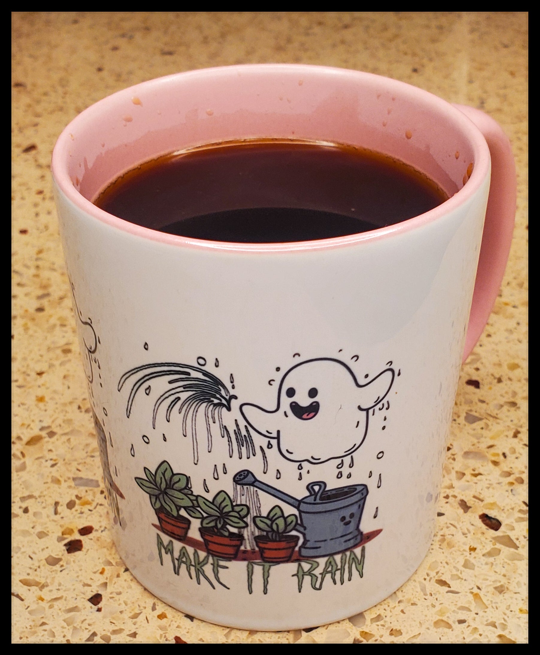 The mug is adorned with a charming illustration featuring a joyful ghost watering potted plants. Above, the phrase "Make it Rain" adds to the whimsy. A pink rim and handle add a soft touch to the design, contrasting nicely with the dark coffee inside.