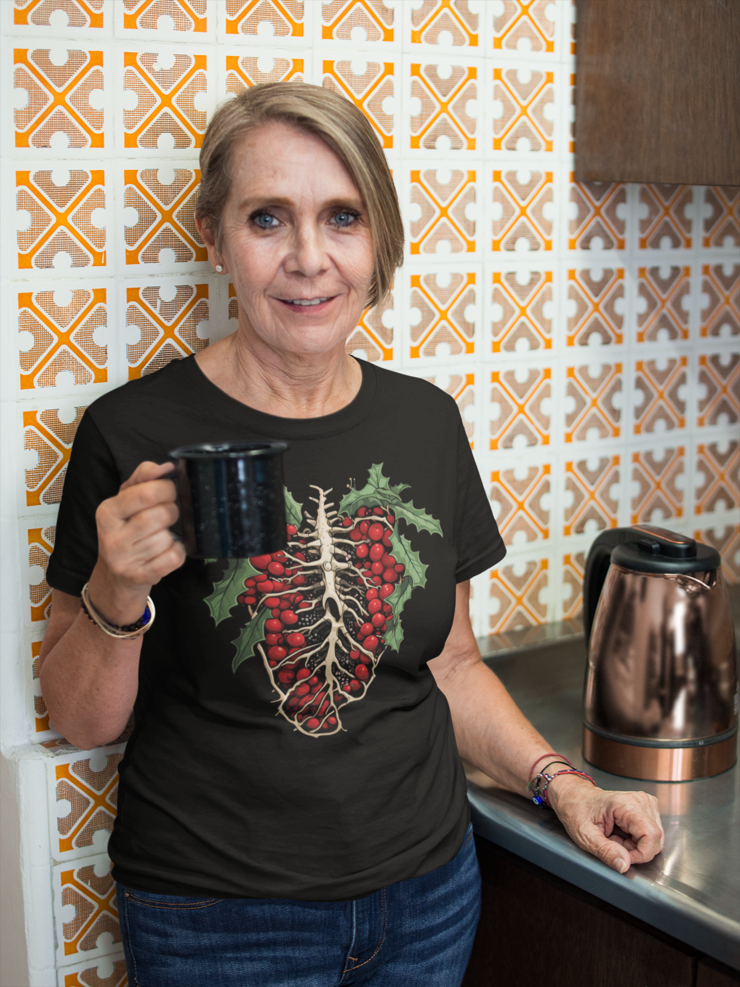 Senior woman in a black t-shirt adorned with a unique holly ribcage design, holding a mug, stands in a kitchen with retro orange patterned tiles, combining homely charm with gothic elements.