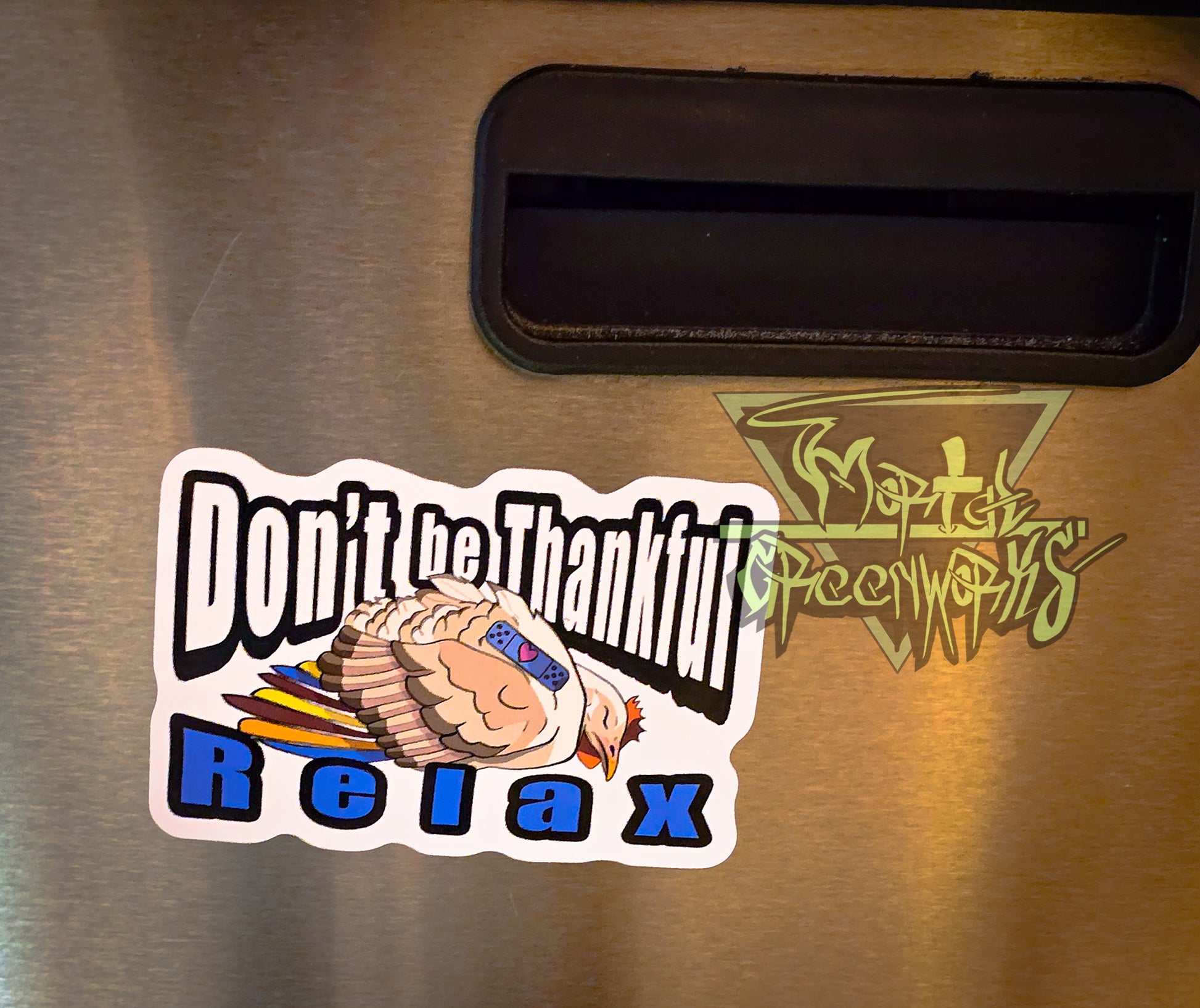 A 4x4" sticker is placed on the side of a shiny stainless steel indoor traschan. The sticker features a beige and brown sleeping turkey with colorful feathers, and a blue sleeping Band-Aid with a heart. In bold Impact text around the turkey reads: "Don't be Thankful, Relax." The Mortal Greenworks logo lingers in the right side of the image.