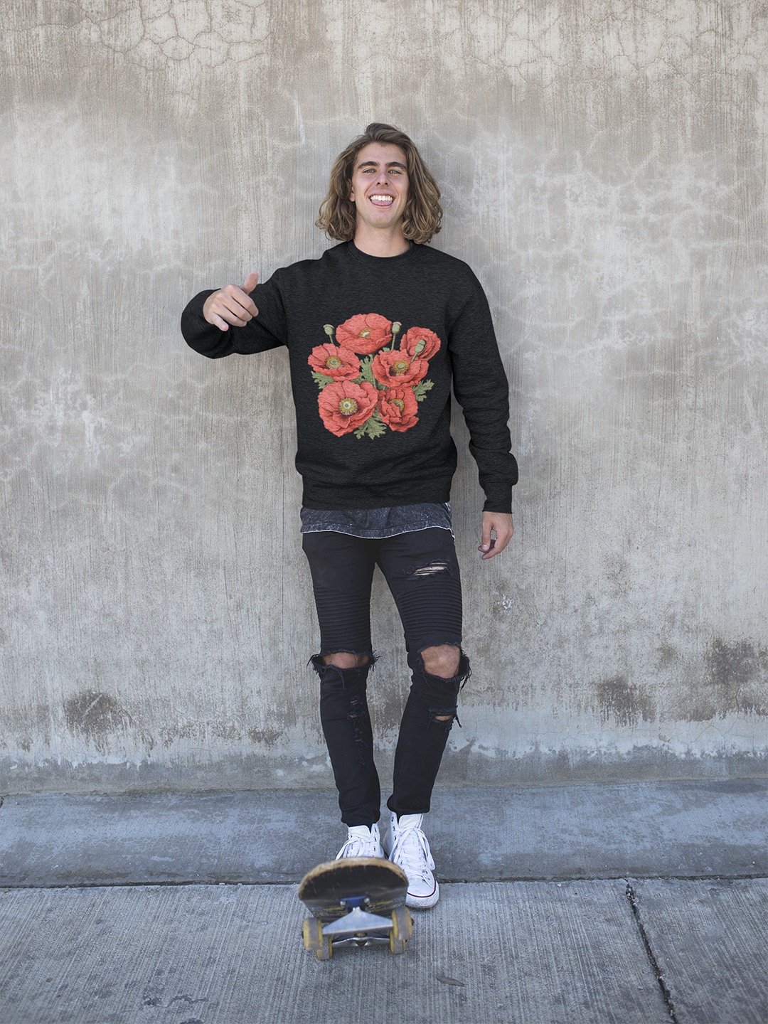 A person stands in front of a textured concrete wall, giving a thumbs-up and smiling. They are wearing a charcoal-colored sweater adorned with a vibrant pattern of red poppies with black centers and green leaves. The individual is also sporting distressed black jeans with pronounced rips at the knees and white sneakers. A skateboard is placed vertically in front of them on the ground, indicating a casual, urban lifestyle.