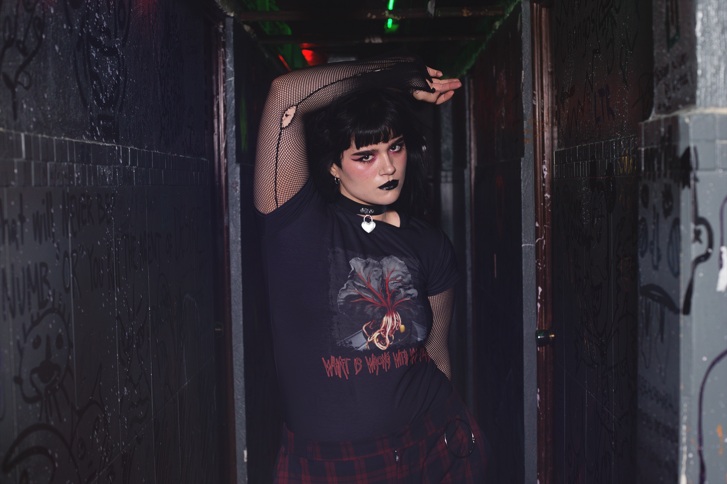 Goth woman in a corridor wears a 'Fiddle Leaf Fig Body Horror' t-shirt, her pose and the setting creating a mood of enigmatic and dark allure.